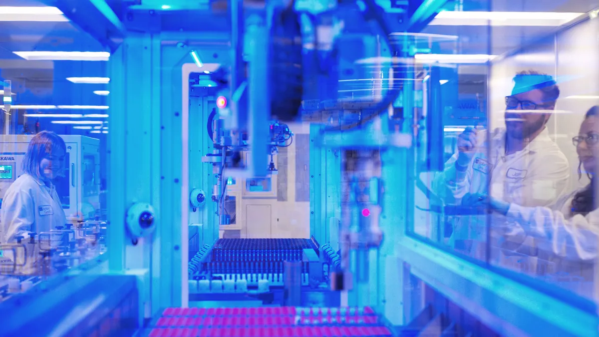 Close up of a robotic arm above several lines of test tubes, with people looking in through a glass window.