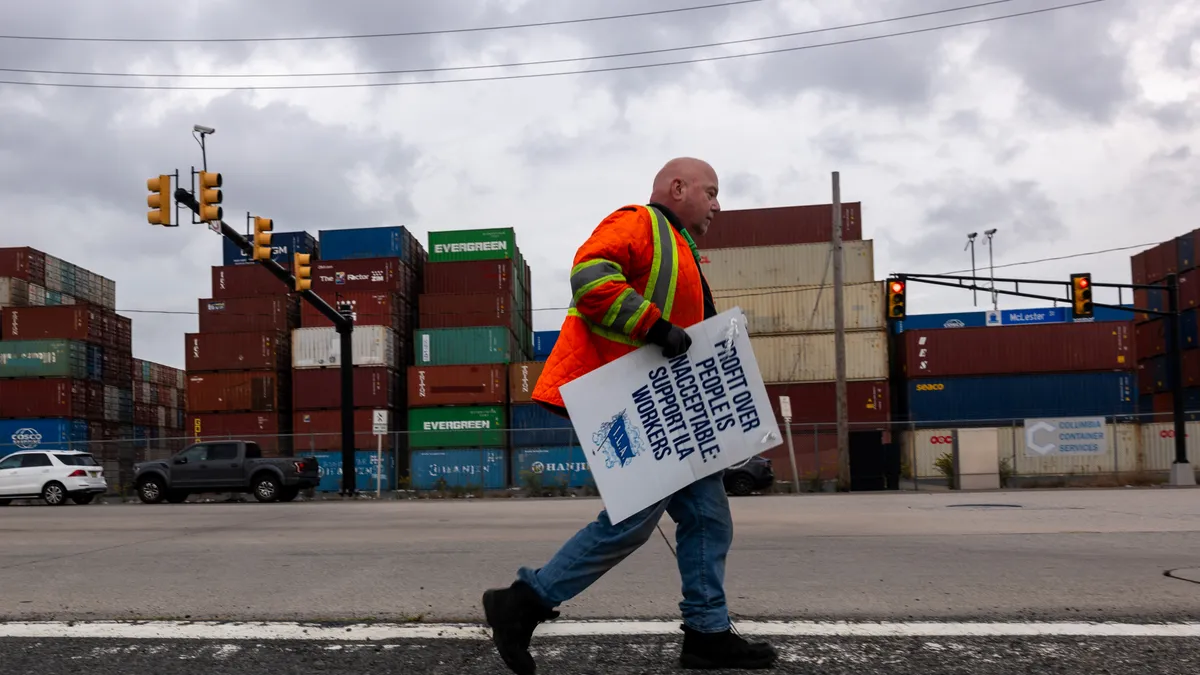 A dockworker on strike walks past shipping containers holding a sign.