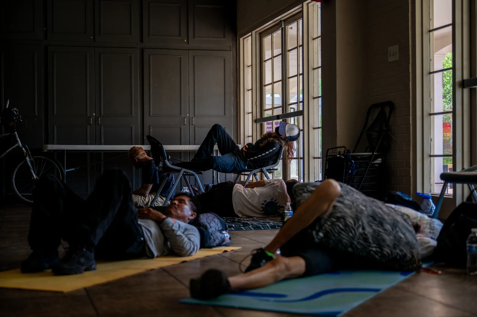 People lay on mats on the ground and in chairs in a dimly lit room.