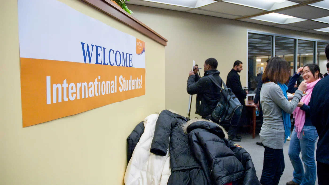 In a large room, a sign in the foreground reads "Welcome International Students!". Multiple groups of students chat amongst themselves in the background.