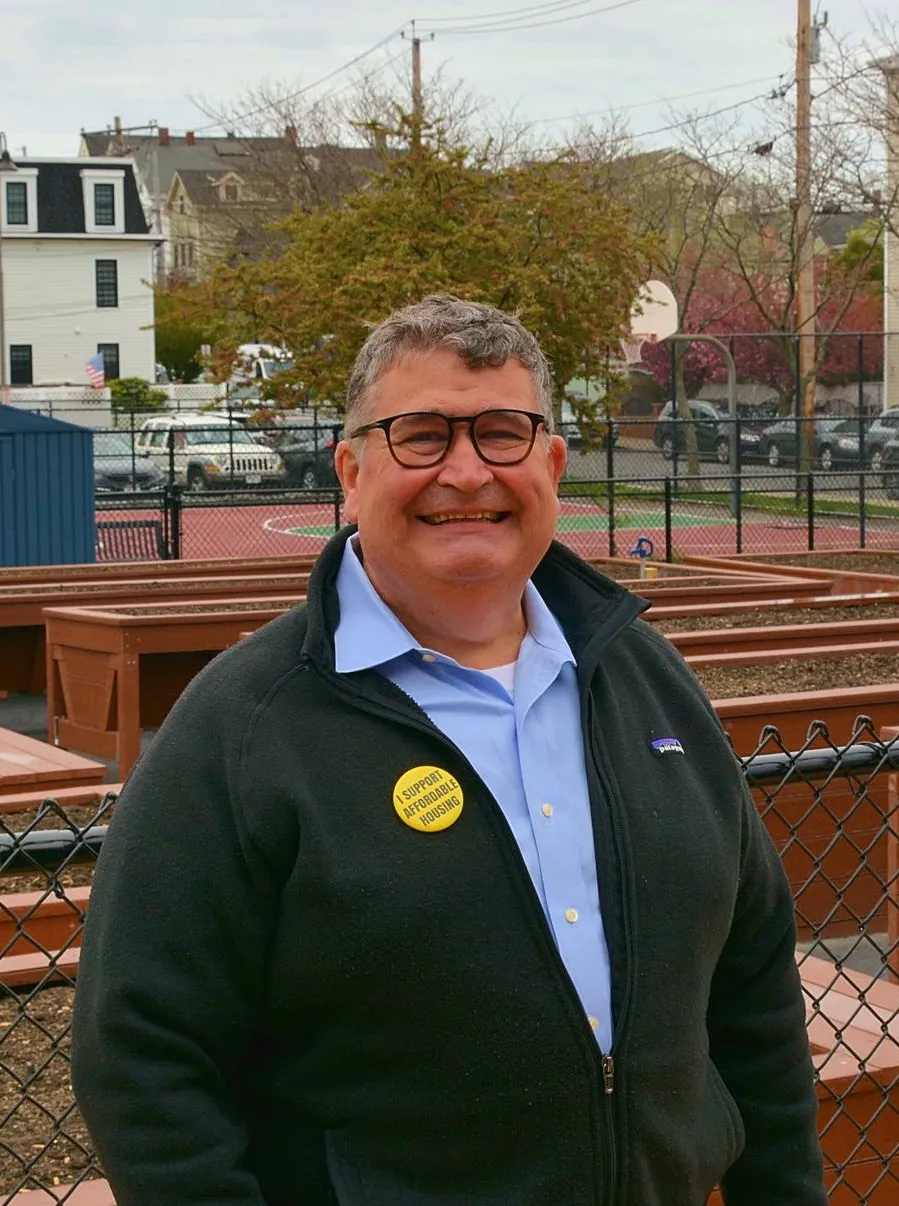 John McQuillan, in glasses and a sweater, stands in a community garden