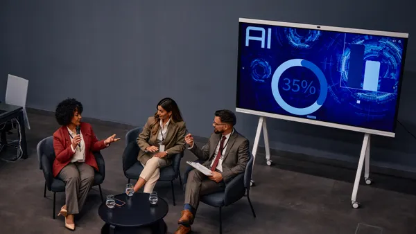 Three businesspeople are sitting in armchairs and discussing artificial intelligence during a summit, with a large screen displaying AI data in the background.