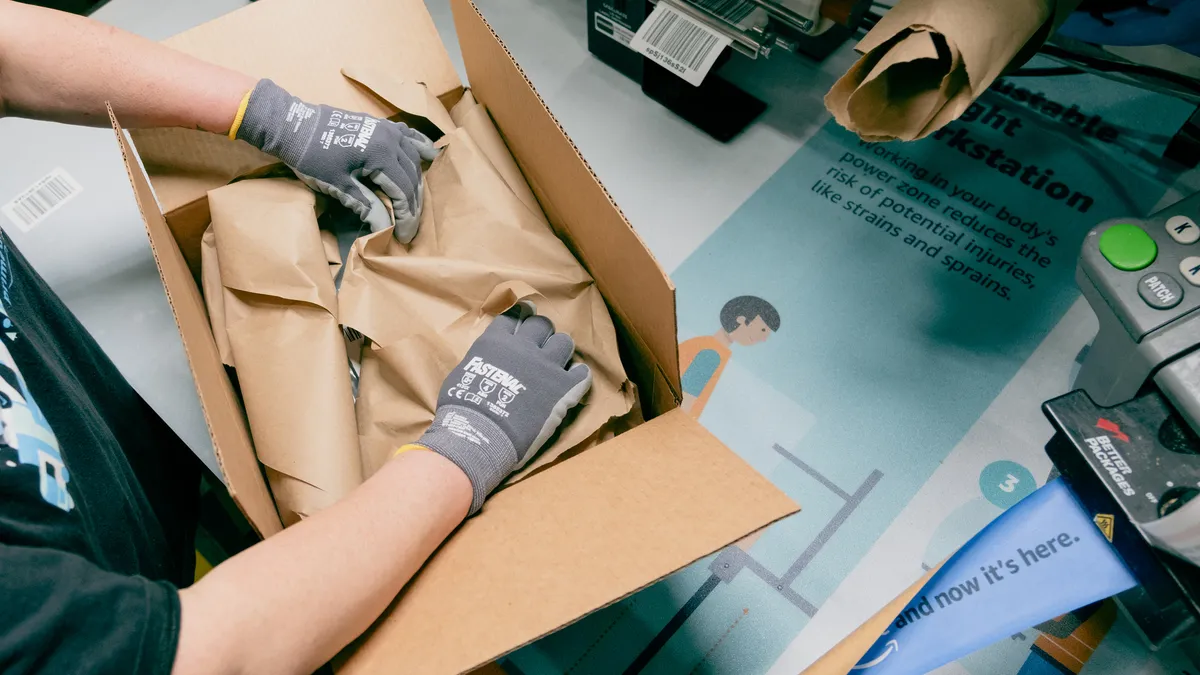 A worker's hands place crumpled paper inside an Amazon delivery box, protecting its contents