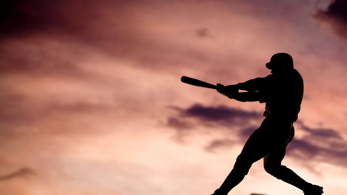 Silhouette shot of baseball player swinging a bat, hitting in the direction of clouds in the sky.