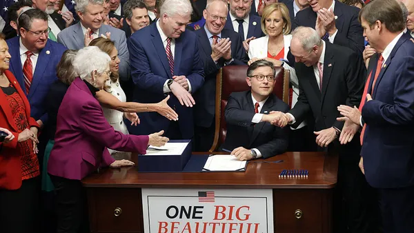One person is sitting at a desk while surrounded behind and on the sides by people with their arms reached out to shake hands. A sign on the desk says "One Big Beautiful Bill Act."