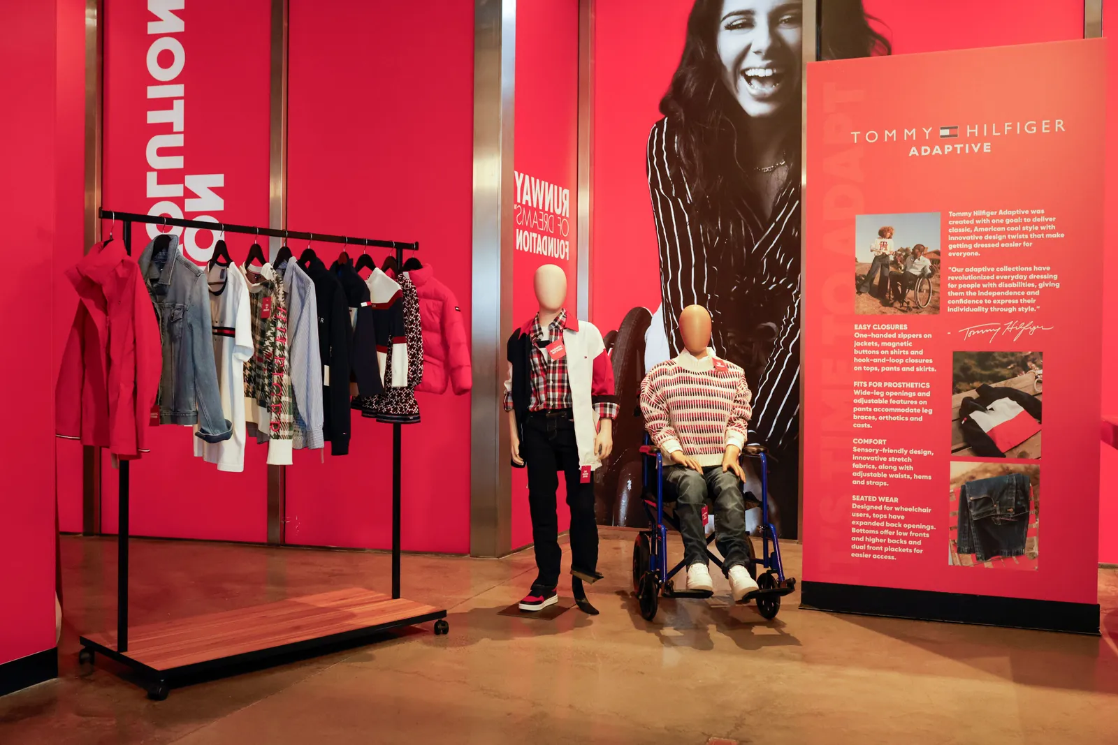 Two mannequins showcase clothes between a rack of clothing and a sign that reads “Tommy Hilfiger Adaptive.” One mannequin has a blade prosthetic leg and the other is posed in a wheelchair.