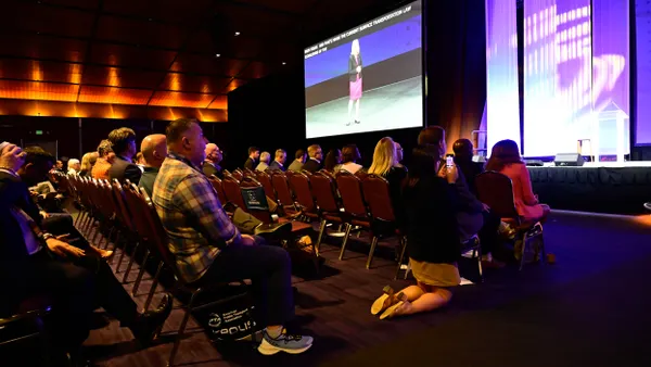 Rows of seated people facing a stage and large screen showing a woman speaking.