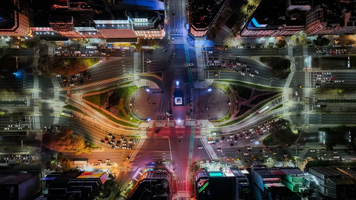 Aerial view of a city intersection at night, illuminated by streetlights and vehicle headlights, showcasing urban activity.