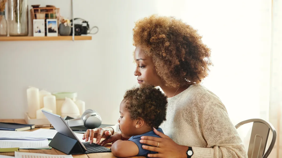 Boy looking at mother using digital tablet. Woman sitting with son at table in kitchen. She is working from home.