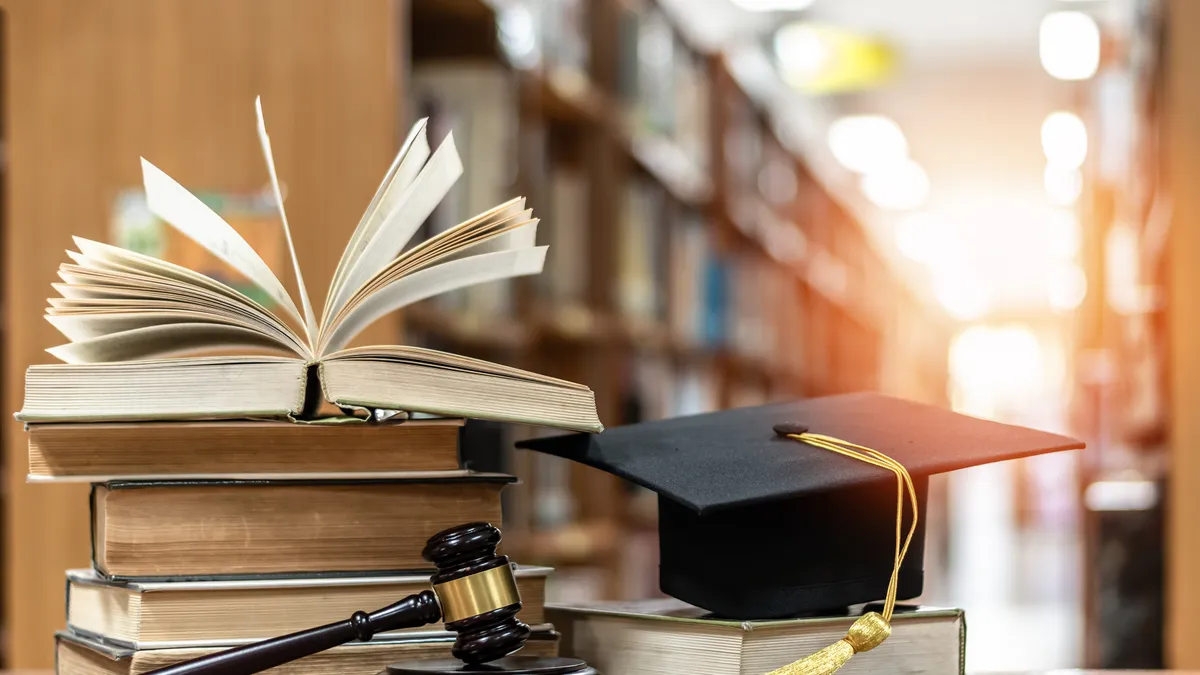 A judge's gavel sits on a desk in front of a stack of books and a graduation cap. In the background are shelves of books.