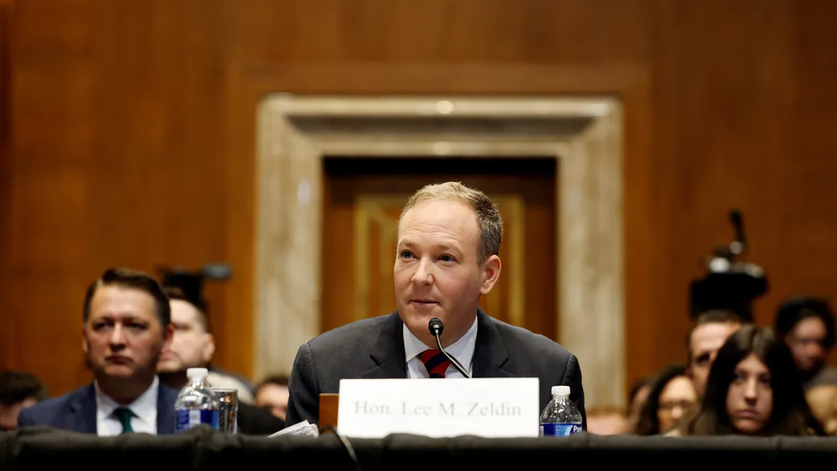 "Hon. Lee M. Zeldin," as a placard states, sits at a table with a microphone in front of a crowd in a hearing room.