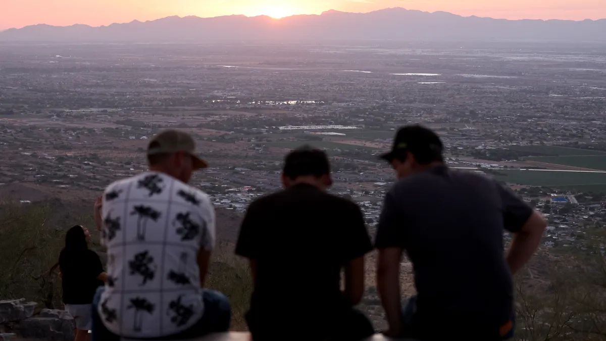 The backs of three people sitting at an overlook over a city and mountains at sunset