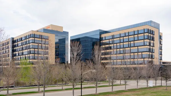 A modern office building with a double glass facade is lined with bare trees in the foreground. A sign reads "Optum" on the building's left side.