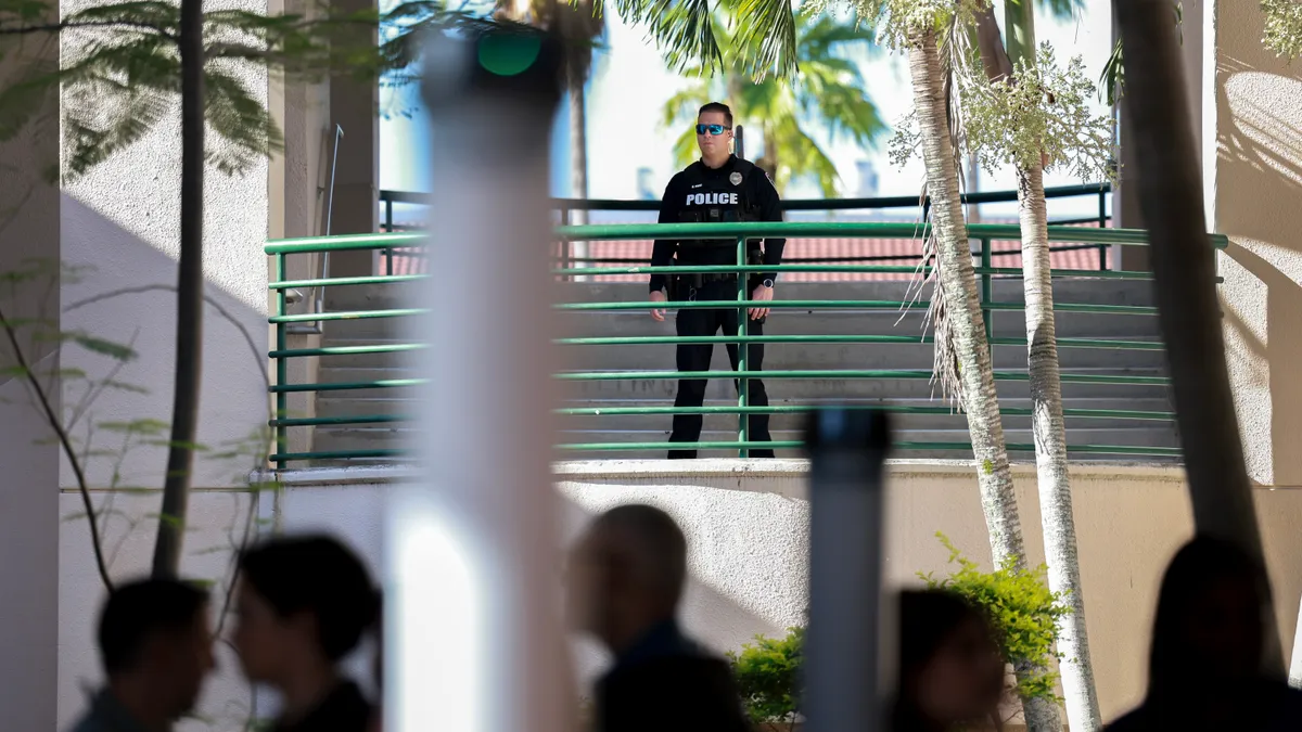 A person in a police uniform stands on a landing above people walking on the ground below who are not in focus.