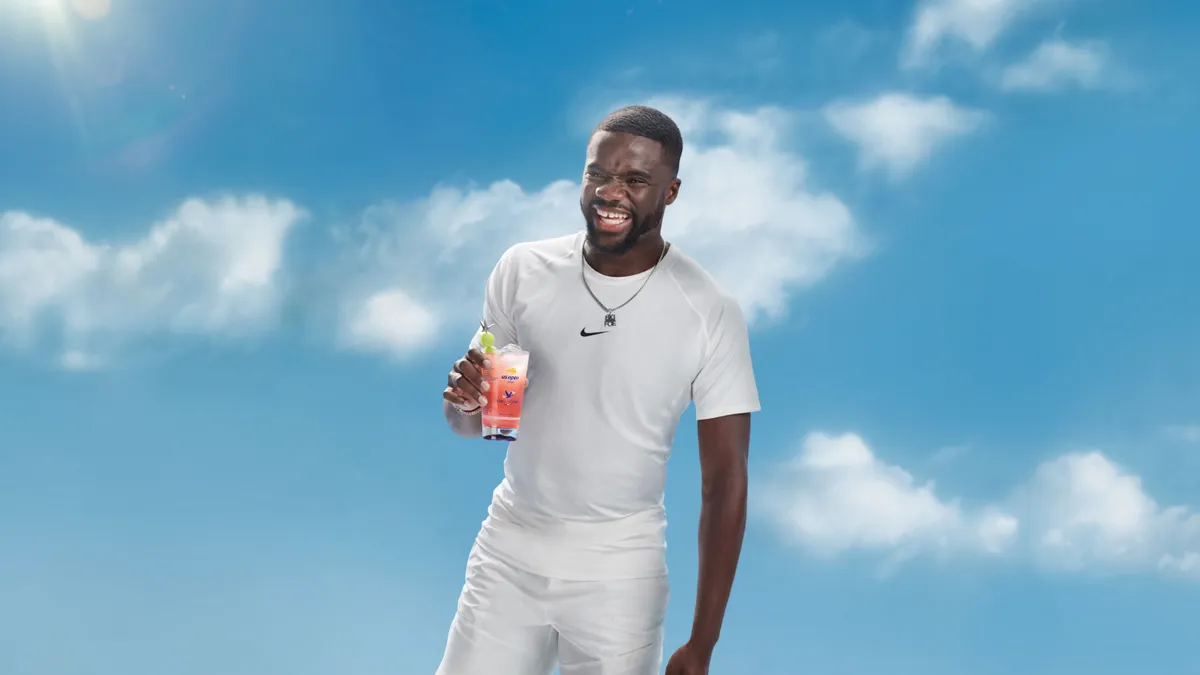 Frances Tiafoe, wearing all white, holds a cocktail against a blue sky background.