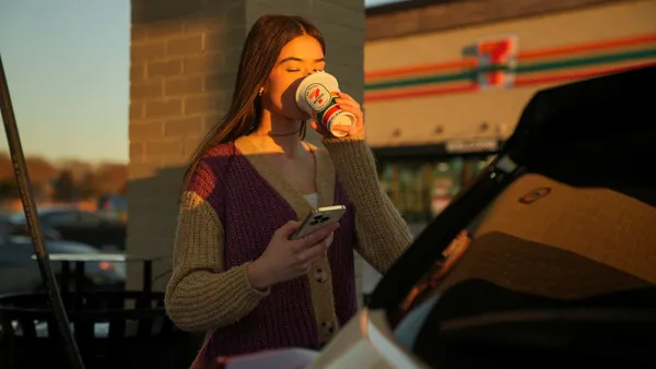 A photo of a person standing outside a store drinking from a disposable coffee cup. The cup and the store behind the person both have a 7-Eleven logo on them.