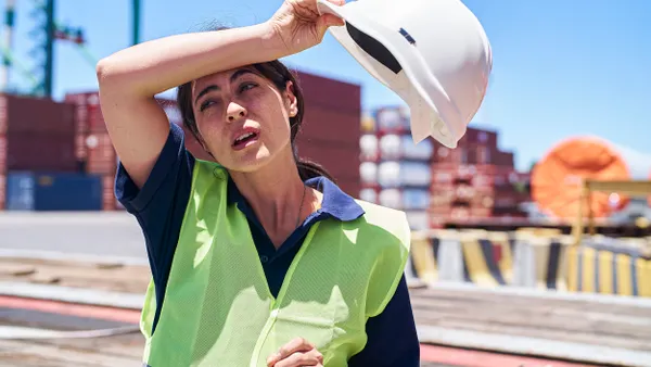 A person wearing a yellow vest holds a white helmet and wipes their brow.