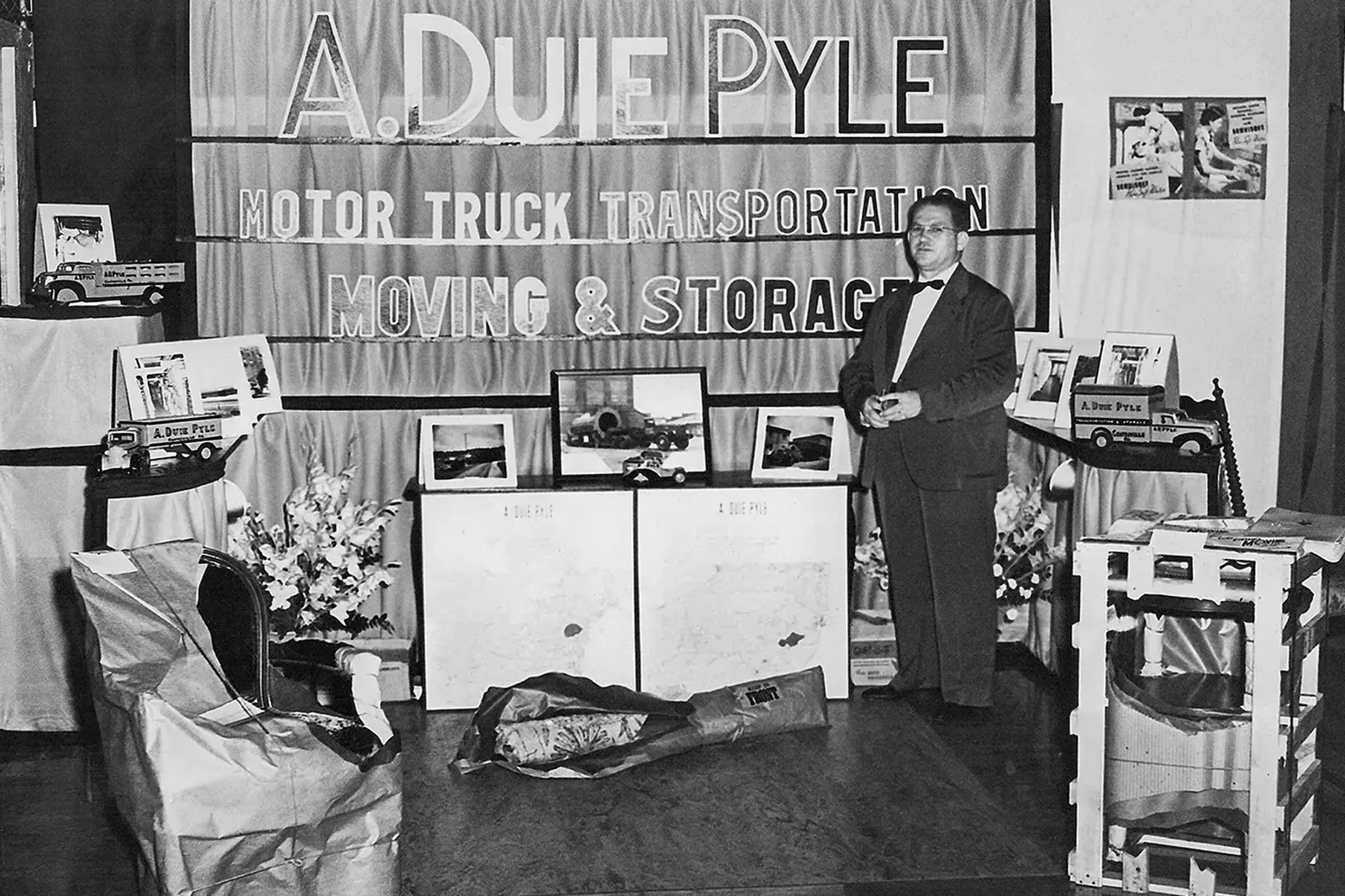 A black n white photo of a person dressed in business attire stands off to the side in a room showcasing trucks and photographs. The backdrop reads, “ A. Duie Pyle, Motor Truck Transportation, Moving &amp; Storage”.