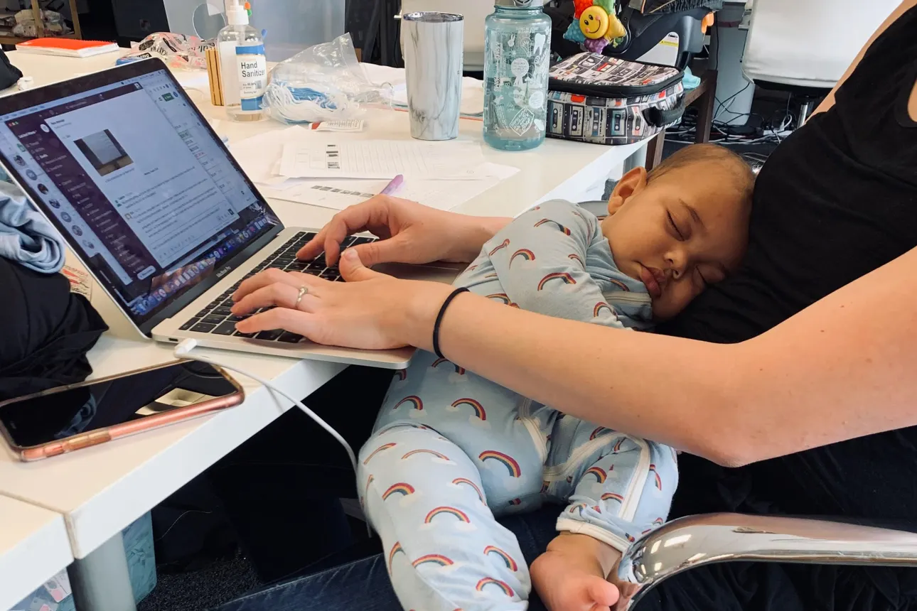 A baby sleeps on its mother's lap as she sits in front of a desk in the office.