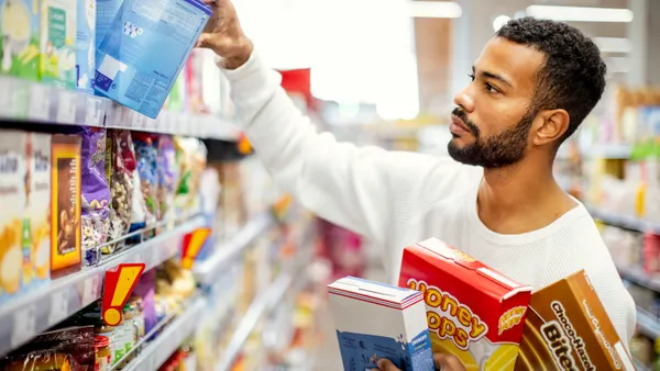 A person holds boxes of food while choosing a cylindrical container from a grocery store shelf.
