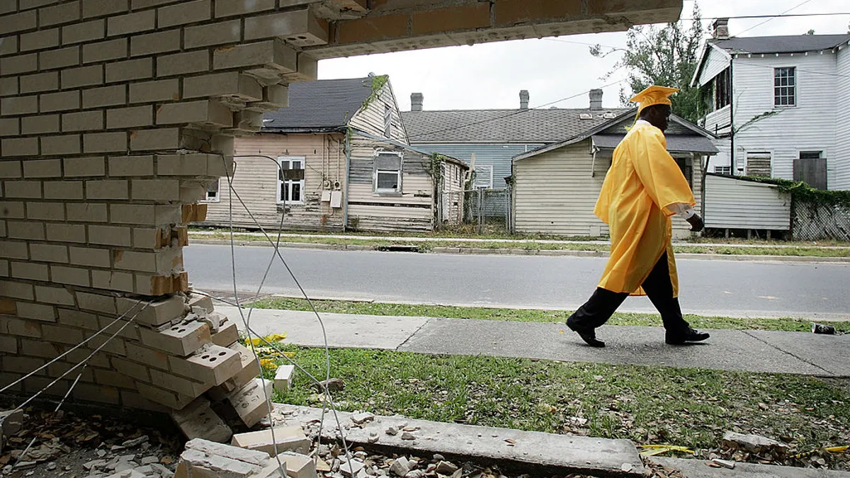 A sideview of a student walking in a graduation cap and gown on a sidewalk silhouetted by a hole in a brick wall.