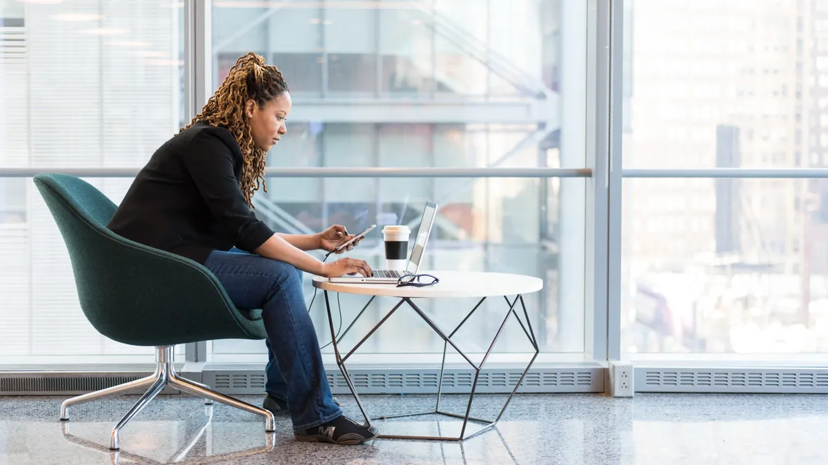 A Black woman sits on blue and gray chair, holding her phone as she scrolls on her computer