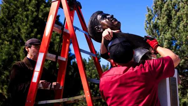 A person wearing a red shirt carries a bronze bust on their shoulder next to a ladder while another person looks on.