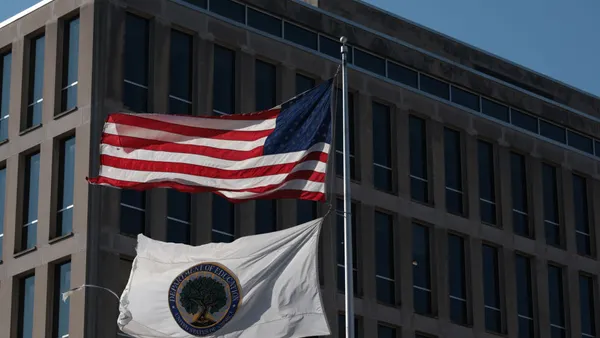 The outside of a building is seen with two flags on a pole flying in front of the building.