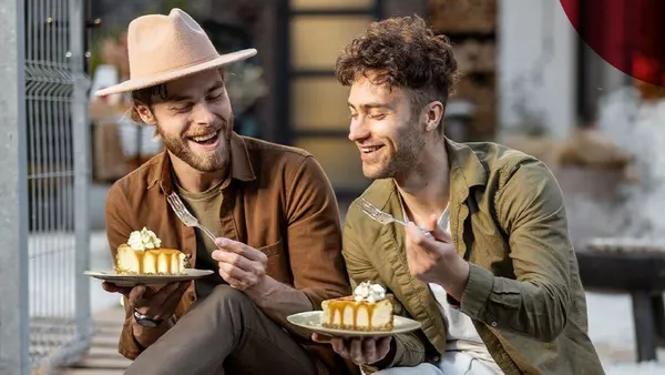 Two men sit outdoors, joyfully eating cake. One wears a hat, the other a green shirt. Both smile and hold plates with caramel-topped desserts.