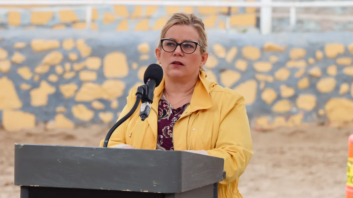 A woman with glasses wearing a yellow jacket speaks into a microphone at a podium.