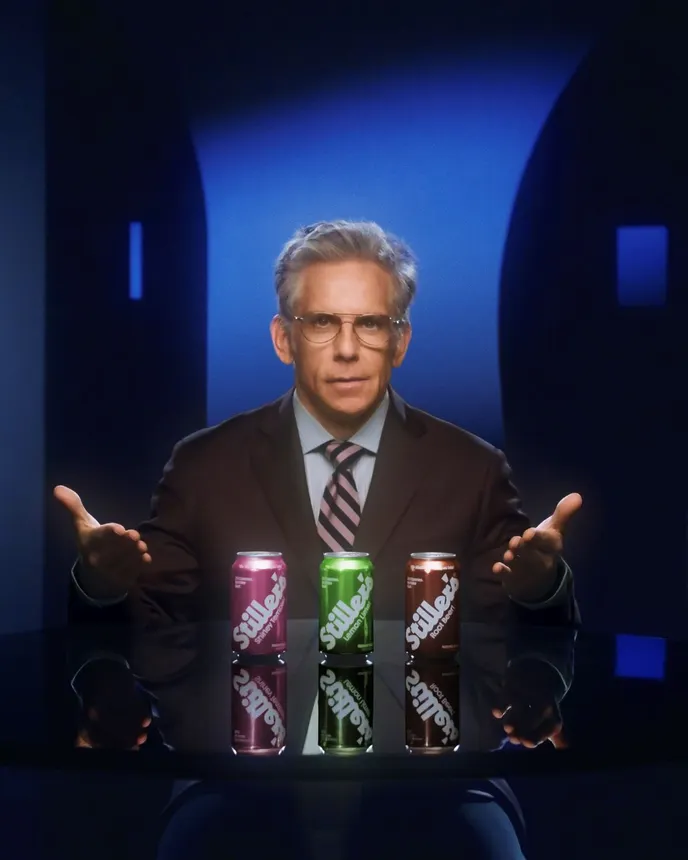 Ben Stiller sits behind a desk on which are three cans of soda.