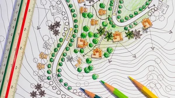 A detailed landscape design plan showing topographic contour lines, orange building illustrations, green trees, and directional arrows. The layout includes winding paths and clustered vegetation. A triangular scale ruler lies on the left, and three colored pencils, colored blue, green, and yellow, rest on the lower right corner of the page.