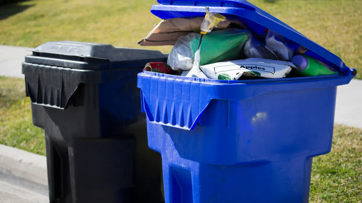 Black trash bin next to blue recycling bin, which has lid propped open