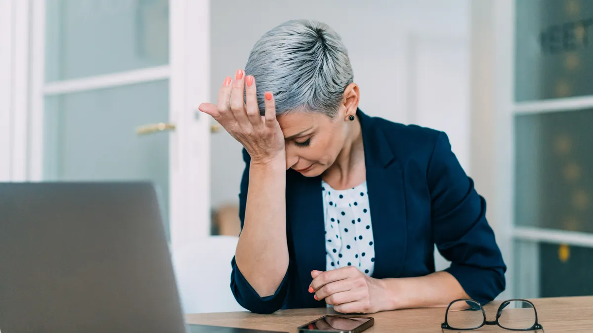 A frustrated businessperson with their head in their hands sitting at a desk in a modern office.