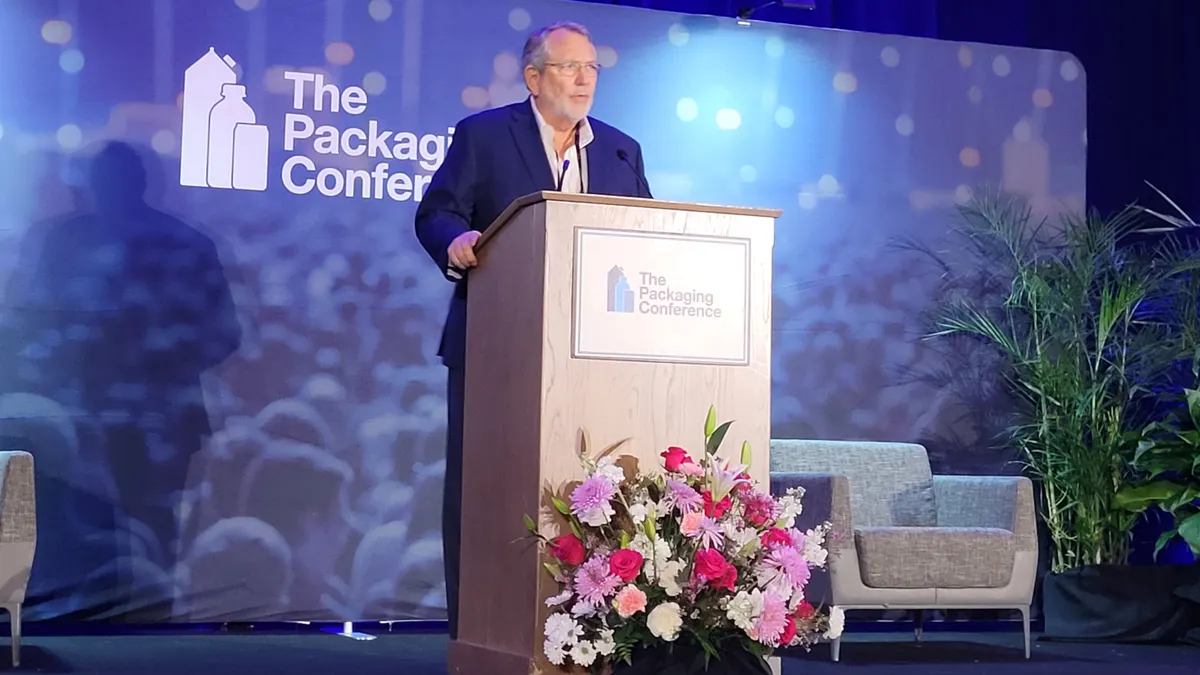 A person stands behind a podium that's in front of a blue background that says The Packaging Conference.