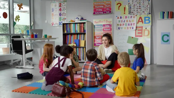 An adult, facing camera, sits on the floor in a classroom. Also on the floor with backs toward camera are students