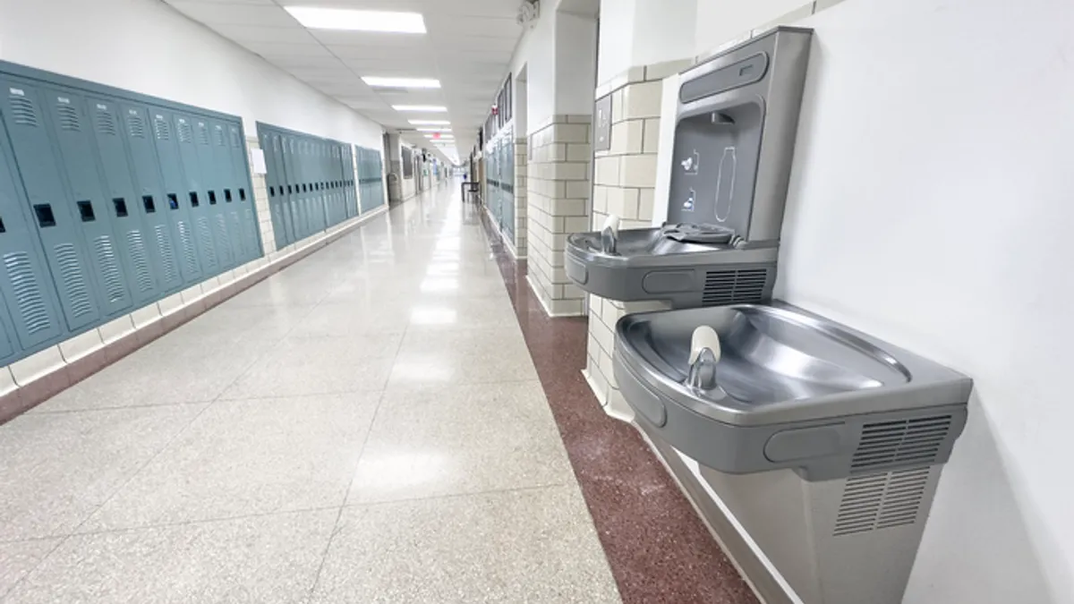 A wall-mounted water fountain is shown in a school hallway. Across the hall are lockers.