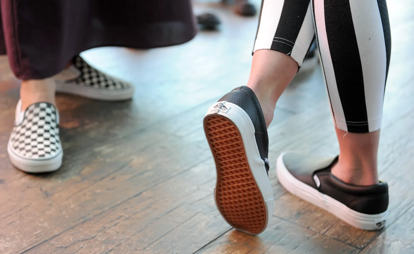 Image shows the ankles and feet of two people wearing new Vans slip-on shoes and standing on a wooden floor