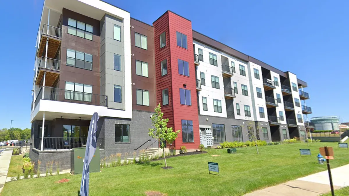 A mid-rise apartment building. Several flags and signs are on the lawn.