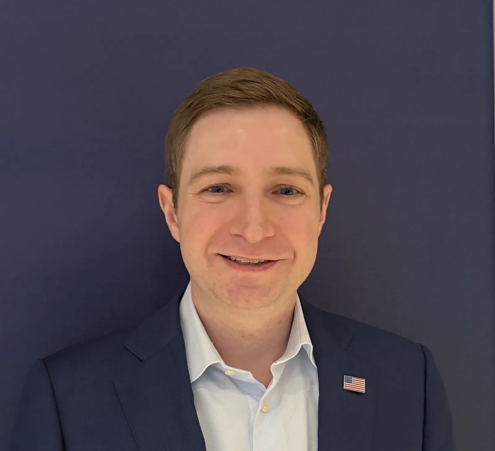 A headshot of a person in a blue suit jack with an American flag pin smiling