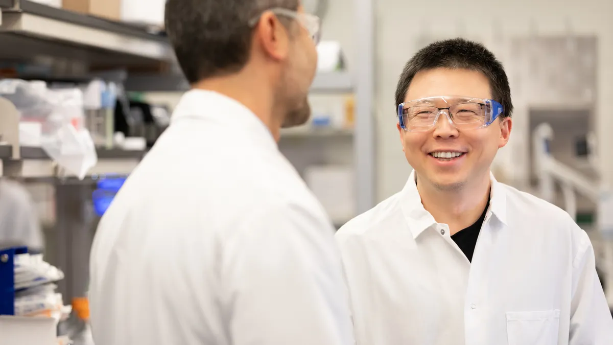A photo of Feng Zhang, founder of Aera Therapeutics, speaking with Akin Akinc, the company's CEO. Both are wearing goggles and lab coats.