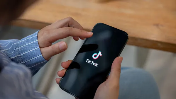 A person holding a smartphone with the TikTok splash screen visible, seated at a table with a hand about to tap the screen.