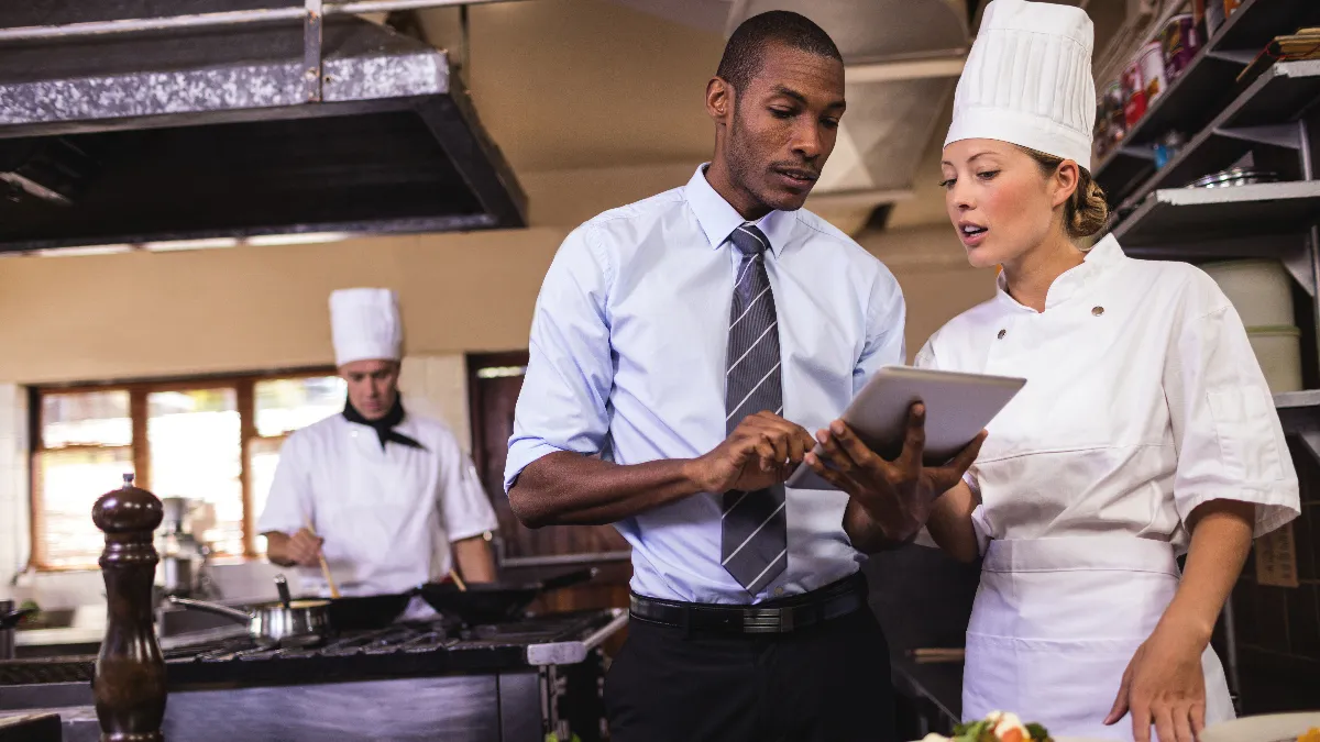 A man and a woman dressed as a chef look at a tablet.