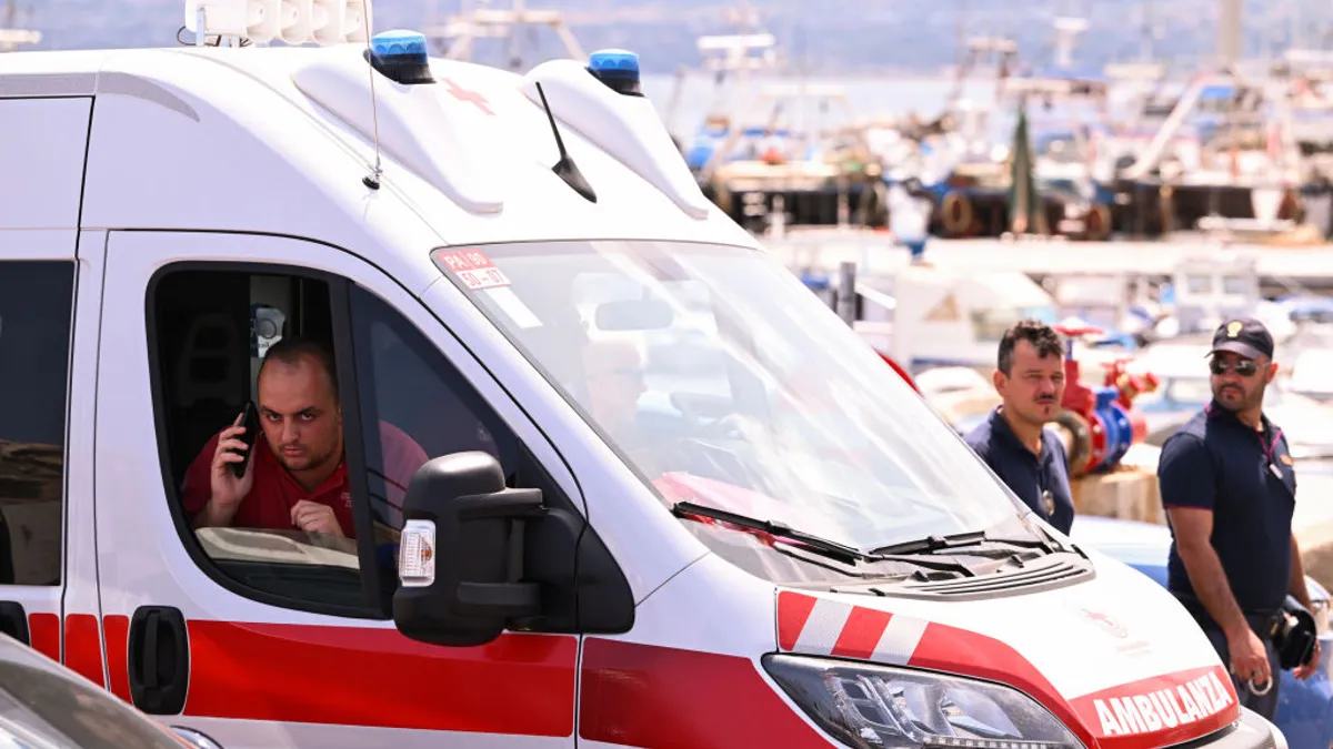 A person talks on a phone inside an ambulance while two others pass by the vehicle, amid recovery efforts for the sunken yacht, Bayesian.