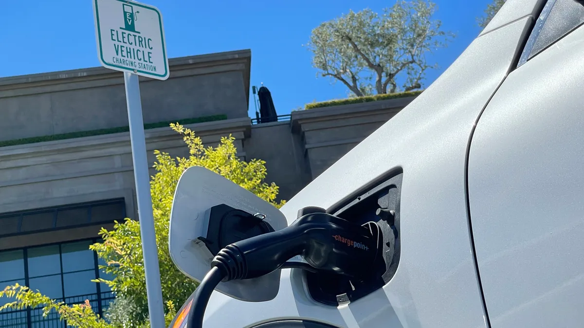 A white electric vehicle, parked by a sign that reads "Electric Vehicle charging station," is seen from a low angle outside the driver's door, plugged in to a charger.