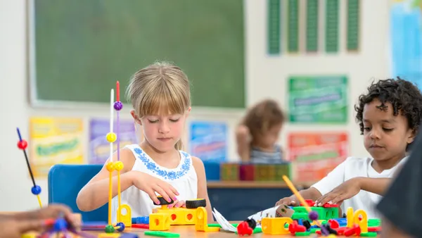 Two young students sit at a desk in a classroom. They are working with colorful blocks and sticks on the desk.