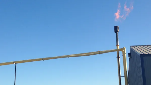 A flare burns gas coming out of a thin metal pipe, part of a system attached to a metal shed.