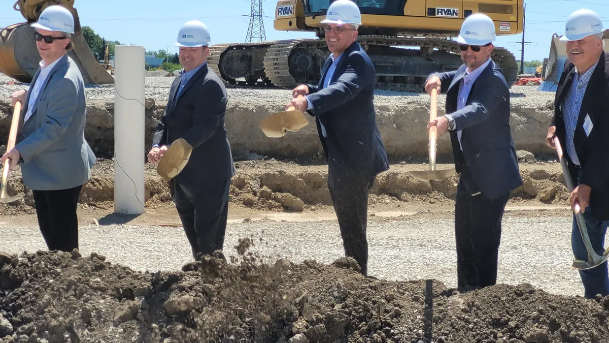 Five people wearing hardhats use shovels to throw dirt during a groundbreaking ceremony.