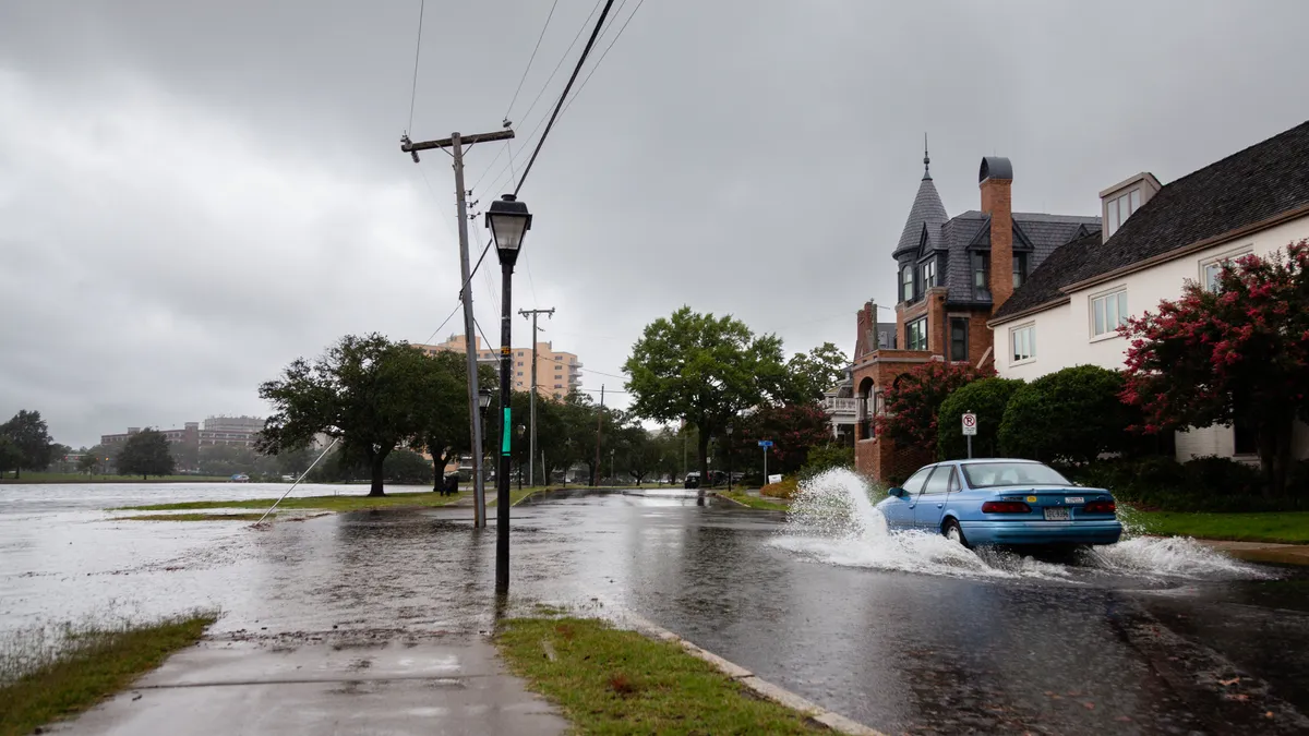 A blue car drives through a flooded street, spraying up water.