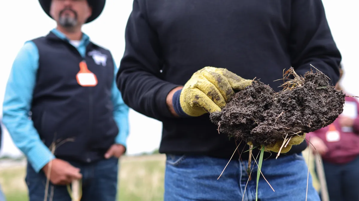 A rancher holds a glob of soil, while another rancher stands behind him.